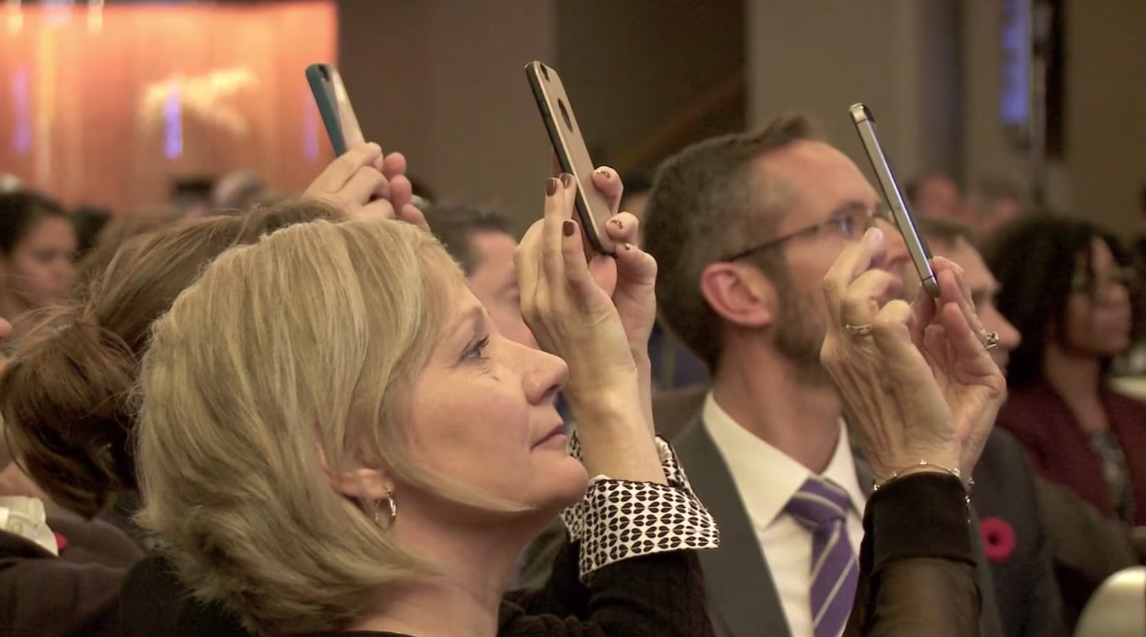 Audience members holding up smartphones to take photos during a formal event.